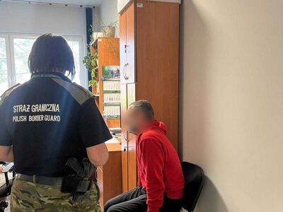 Na zdjęciu mężczyzna w czerwonej bluzie, siedzi na krześle przy ścianie. Obok niego kobieta, prawdopodobnie funkcjonariuszka Straży granicznej ubrana w czarny T-shirt i zielone spodnie, Na plecach napis Straż Graniczna/ Polish Border Guard. W tle meble biurowe.