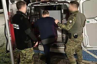 Dwóch funkcjonariuszy, jeden w czarnym mundurze z napisem w jasnym kolorze na plecach (częściowo zasłoniętym) i drugi w mundurze kamuflażowym, wprowadza mężczyznę ubranego na ciemno do tyłu furgonetki. Tył furgonetki jest otwarty i widoczna jest metalowa krata oddzielająca przestrzeń ładunkową.