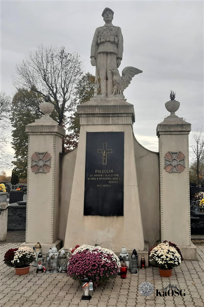 Pomnik na cmentarzy w radłowie, pod nim leża chryzantemy i w centralnym miejscu znicz  z biało - czerwoną tasiemką i logiem Karpackiego Oddziału Straży Granicznej