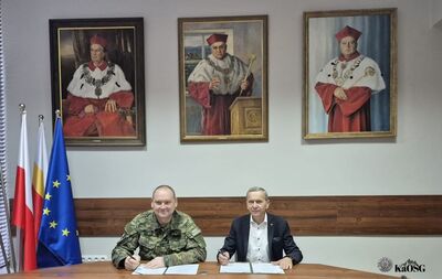 Dwóch mężczyzn, jeden w mundurze wojskowym, drugi w garniturze, siedzi za dużym drewnianym stołem i podpisuje dokumenty. W tle, na ścianie, wiszą trzy portrety przedstawiające historyczne postacie w strojach akademickich, a obok stoją flagi Polski i Unii Europejskiej.