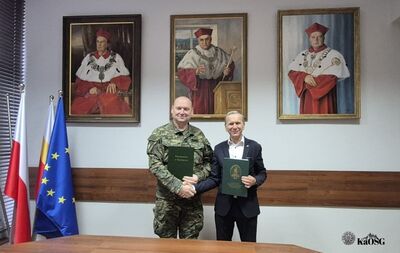 Dwóch mężczyzn stoi za drewnianym stołem i ściska sobie dłonie. Mężczyzna po lewej w mundurze trzyma zielony dokument z napisem &quot;Straż Graniczna&quot;, a mężczyzna po prawej, w garniturze, trzyma podobny zielony folder. Za nimi wiszą trzy portrety postaci w czerwonych togach i biretach.