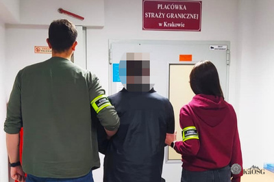 Na zdjęciu widać trzy osoby stojące tyłem. Osoba w środku trzymana jest przez dwie pozostałe za ręce jako osoba zatrzymana. Po lewej stronie zdjęcia stoi mężczyzna ubrany w zieloną kurtkę, rękaw na lewej ręce ma podwinięty do łokcia, na nadgarstku ma czarny zegarek, na drugiej ręce ma odblaskową opaskę z napisem Straż Graniczna. W środku stoi mężczyzna ubrany w granatową kurtkę. Po prawej stronie zdjęcia stoi kobieta z długimi ciemnymi włosami ubrana w bordową bluzę z kapturem na lewej ręce ma odblaskową opaskę z napisem Straż Graniczna, na nadgarstku założony ma zegarek z białym paskiem.