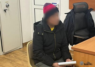 Na zdjęciu mężczyzna siedzący w pomieszczeniu służbowym. Jest ubrany w czarną kurtkę zapinaną na zamek i kolorową czapkę. W ręku trzyma zwinięte w rulon kartki papieru. W tle widoczne drzwi wejściowej, w prawym rogu znajduje się fragment biurka i krzesło biurowe.