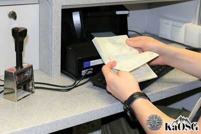 zbliżenie na dłonie funkcjonariusza, który trzyma w nich otwarty paszport, na biurku leży stempel kontrolerski i klawiatura