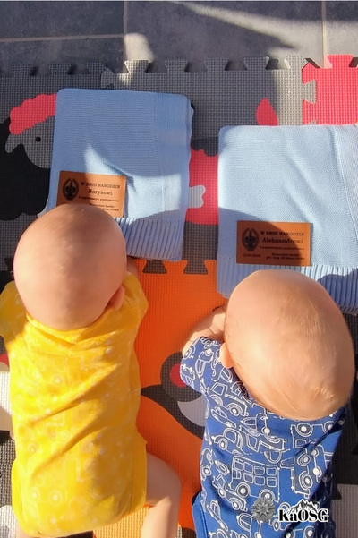 dwoje niemowlaków leży na brzuszku i patrzy na niebeskie kocyki złożone w kostkę, otrzymane od komendanta kaosg