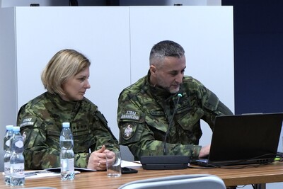 Dwie osoby siedzą przy stole ubrani są w mundury zielone. Mężczyzna po prawej stronie referuje. Tłem jest wyświetlony slajd na komputerze.