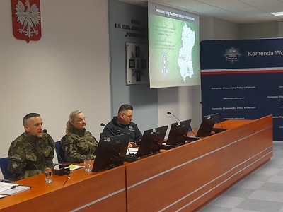 Trzy osoby w mundurach siedzą przy stole. W tle jest wielki monitor na którym wyświetlane są dane.