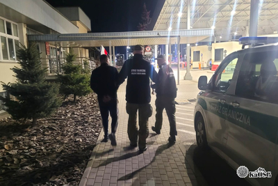 Na zdjęciu widać trzy osoby, które idą w stronę dużego budynku. Po prawej stronie widać oznakowany samochód Straży Granicznej.