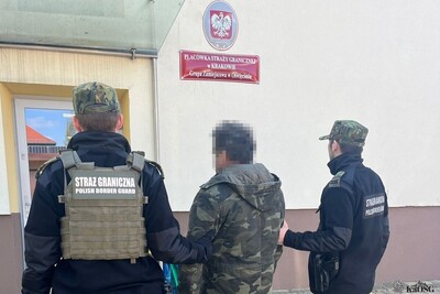 Na zdjęciu widać trzy osoby stojące przed budynkiem państwowym. Dwie z nich ubrane są w mundury. Prowadzą zatrzymaną osobę do budynku.