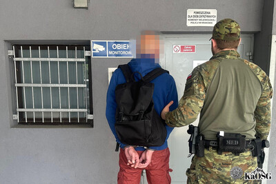 Zatrzymany mężczyzna stoi tyłem w asyście funkcjonariusza sg przed wejściem do pomieszczeń dla osób zatrzymanych w komendzie wojewódzkiej policji w krakowie, na rękach ma kajdanki a jego głowa jest zapikselizowana