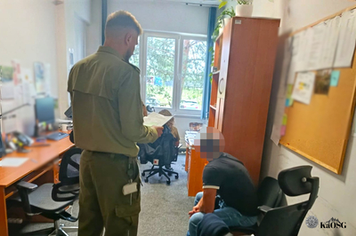 Na zdjęciu widać dwie osoby w pomieszczeniu. Jedna po lewej stoi, a po prawej stronie widać osobę która siedzi.