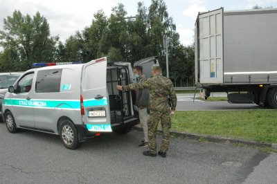 Zatrzymany cudzoziemiec  prowadzony jest przez funkcjonariusza SG do samochodu typu więźniarka.