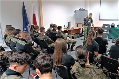 Uczniowie w mundurach siedzą w sali i słuchają pogadanki. Przed nimi stoi funkcjonariusz Straży Granicznej - przewodnik psa służbowego opowiadający o swojej służbie.