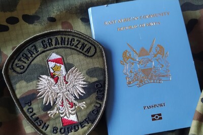 Służbowo z Kenii przez Hiszpanie do Polski emblemat straży granicznej z napisem straż graniczna polish border guard na środku emblematu znajduję się orzeł biały a za nim słupek graniczny białoczerwony. obok emblematu znajduje się paszport Republiki Kenii w kolorze niebieskim. emblematy oraz paszport znajdują się na materiale o wzorze  moro
