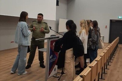 Sala wykładowa. Funkcjonariusz Straży Granicznej stoi z grupą studentów i odpowiada na ich pytania.