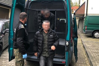 Zatrzymanych trzech cudzoziemców pod nadzorem funkcjonariusza Straży granicznej wysiada z pojazdu służbowego oznakowanego typu więźniarka. Cudzoziemcy mają zapikselizowane twarze.