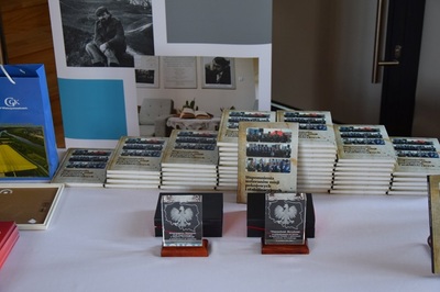 Na stole leży kilka stosów książek pt. Wspomnienia weteranów misji pokojowych i stabilizacyjnych z powiatu tarnowskiego. Przed nimi stoją dwie szklane statuetki z wizerunkiem orła.