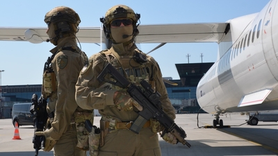 W tle samolot pasażerski stojący na płycie lotniska. Przed nim stoi dwóch zamaskowanych i uzbrojonych funkcjonariuszy Zespołu Interwencji Specjalnych z podkrakowskiego lotniska.