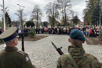 Nowy Sącz. Plac przed pomnikiem marszałka Józefa Piłsudskiego. Tyłem stoją funkcjonariusze pełniący wartę honorową. Przed nimi duża grupa mieszkańców Nowego Sącza z kwiatami i biało-czerwonymi flagami.