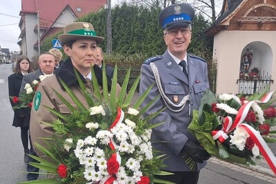 Funkcjonariuszka SG z biało-czerwonym wieńcem idzie w kolumnie uczestników uroczystości rocznicowych w Czarnym Dunajcu. Towarzyszy jej policjant z wiązanką biało-czerwonych kwiatów. Po prawej kapliczka.