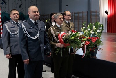 Delegacja służb mundurowych niesie wiązankę biało-czerwonych kwiatów.