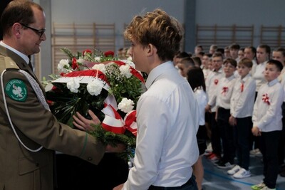 Delegacja Karpackiego Oddziału Straży Granicznej przekazuje uczniowi wiązankę biało-czerwonych kwiatów. W tle uczniowie ubrani w białe koszule z biało-czerwonymi kotylionami.