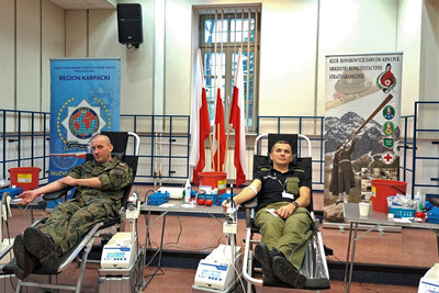 Oddali honorowo krew w Karpackim Oddziale SG Funkcjonariusz Straży Granicznej oraz żołnierz oddający krew. na drugim planie flagi Polski oraz banery informacyjne.