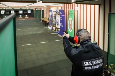 Pomieszczenie w strzelnicy. Funkcjonariusz Straży Granicznej w słuchawkach ochronnych oddaje strzał z pistoletu do tarczy.