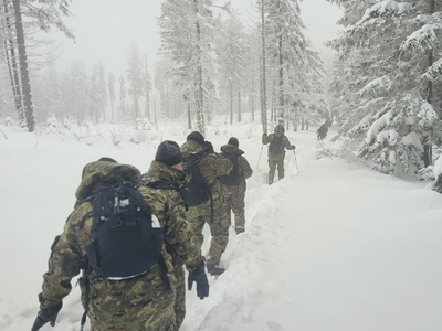 Zimowa sceneria, bardzo dużo śniegu. Grupa umundurowanych funkcjonariuszy Straży Granicznej niosąca plecaki idzie pod górę szlakiem turystycznym.