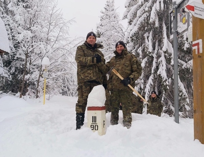 Zimowa sceneria, bardzo dużo śniegu. Generał Jopek i Generał Laciuga stoją przy znaku granicznym III/90 uściskują sobie dłoń. Generał Laciuga trzyma w ręku ciupagę góralską.