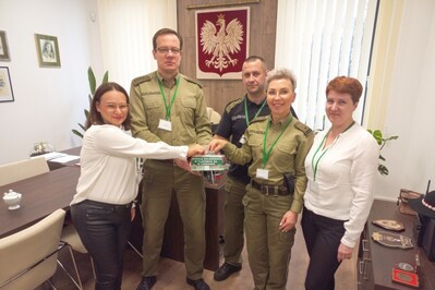 Pomieszczenie komendanta placówki SG w Tarnowie. Dwóch pracowników i trzech funkcjonariuszy wspólnie trzyma puszkę na zbiórkę koleżeńską i wrzucają pieniądze.