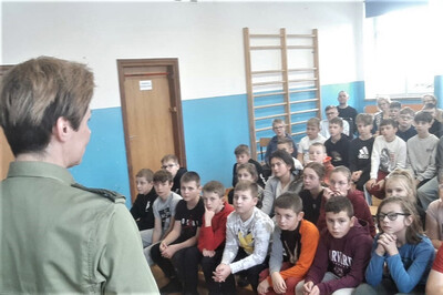 Sala gimnastyczna szkoły. Funkcjonariuszka stoi przed kilkudziesięcioosobowa grupą młodzieży, prowadzi prelekcję na temat służby w Straży Granicznej.