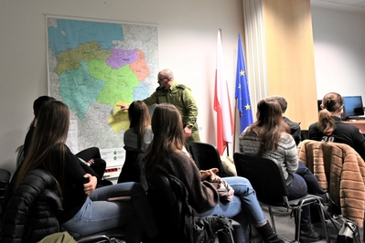 Sala szkoleniowa w siedzibie Placówki SG w Krakowie-Balicach. Funkcjonariusz SG stoi przy mapie Polski. Opowiada studentom o służbie w Straży Granicznej.