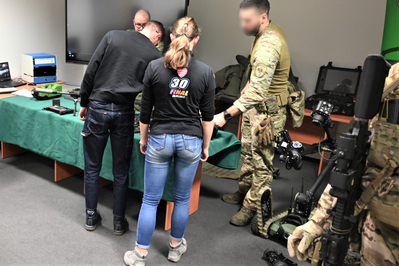 Sala szkoleniowa w siedzibie Placówki SG w Krakowie-Balicach. Funkcjonariusze SG stoją przy stole na którym wystawiony jest sprzęt służbowy używany na co dzień przez funkcjonariuszy SG. Młodzi ludzie oglądają broń i wyposażenie SG.