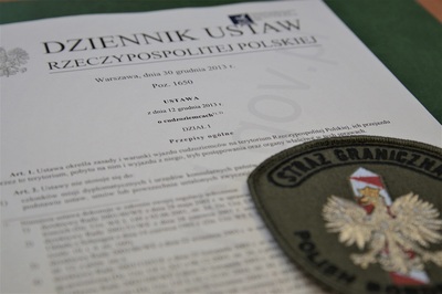 Kolejni cudzoziemcy nielegalnie w Polsce emblemat straży granicznej znajdujący się na papierowej wersji ustawy o cudzoziemcach. całość położona na biurku.