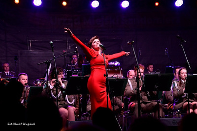 Sala koncertowa. Solistka orkiestry ubrana w czerwoną wieczorowa suknie śpiewa na scenie. Za nią muzycy orkiestry reprezentacyjnej straży granicznej
