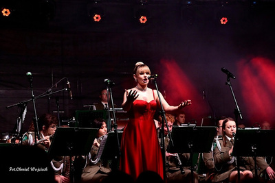 Sala koncertowa. Solistka orkiestry ubrana w czerwona wieczorową suknię  śpiewa na scenie. Za nią muzycy orkiestry reprezentacyjnej straży granicznej