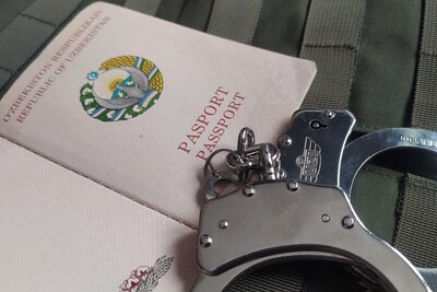 Na kamizelce taktycznej w kolorze zielonym leży otwarty paszport obywatela uzbekistanu, a na nim w dolnym rogu leżą kajdanki.