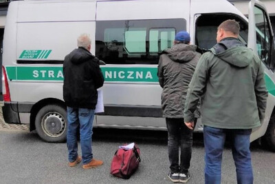 Zatrzymani obywatele turcji stoją przed otwartym radiowozem straży granicznej. między mężczyznami leży torba podróżna