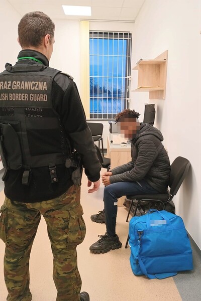 Nielegalny pobyt obywateli RPA oraz Algierii zatrzymany cudzoziemiec siedzi w pomieszczeniu służbowym straży granicznej w obecności funkcjonariusza straży granicznej. obok krzesła na którym siedzi zatrzymany mężczyzna znajduje się jego torba z rzeczami.