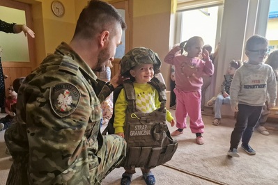 Dziecko ubrane w hełm oraz kamizelkę kuloodporną , uśmiechające się do funkcjonariusza straży granicznej, który je ubierał. na drugim planie inne zebrane dzieci.