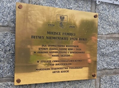 tabliczka informacyjna z napisem miejsce pamięci bitwy niemeńskiej 1920 roku. opis z jakiej okazji  została ufundowana tabliczka. napisy w kolorze czarnym na złotym tle.