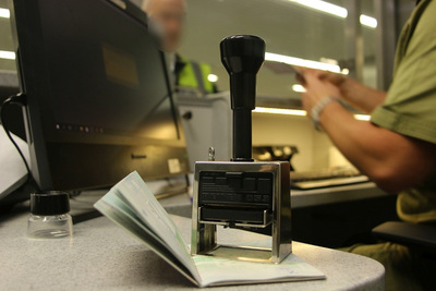 Na biurku leży paszport,, na nim położony stempel kontrolerski. w tle funkcjonariusz sg sprawdzający dokument a przed okienkiem kabiny stoi podróżny