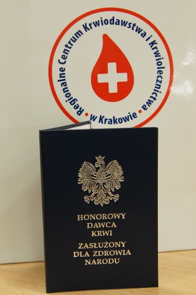 granatowa książeczka Honorowy Dawca Krwi - Zasłużony dla Zdrowia Narodu na tle logotypu regionalnego centrum krwiodawstwa i krwiolecznictwa w krakowie