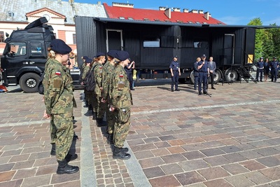 oficjalne rozpoczęcie wydarzenia. przemawia funkcjonariusz policji. po jego lewej stronie stoją w dwuszeregu uczniowie szkoły o profilu mundurowym ubrani w umundurowanie moro. w tle stoi esape room w kolorze czarnym.