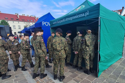 młodzież w umundurowaniu moro, która stoi przy stoisku promocyjnym karpackiego oddziału straży granicznej. stoisko jest w kolorze zielonym z napisem karpacki oddział straży granicznej. nad napisem znajduje się logo oddziału.