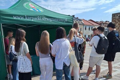 młodzież która stoi przy stoisku promocyjnym karpackiego oddziału straży granicznej. stoisko jest w kolorze zielonym z napisem karpacki oddział straży granicznej. nad napisem znajduje się logo oddziału.