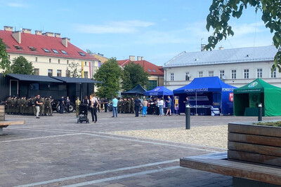 ogólne zdjęcie na wydarzenie zorganizowane na placu w Olkuszu. widoczny jest pojazd tak zwany esape room oraz funkcjonariusz policji przemawiający do zebranych na wydarzeniu osób. w dwuszeregu stoją uczniowie w mundurach moro ze szkoły o profilu mundurowym.