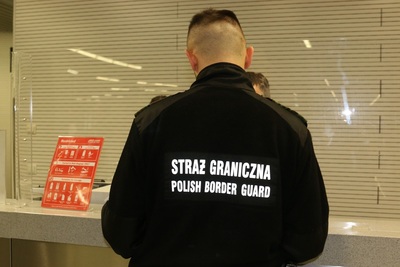 funkcjonariusz straży granicznej stojący przy okienku w porcie lotniczym. jest obrócony tyłem. na czarnym polarze służbowym widoczny jest biały napis Straż graniczna oraz Polish border guard.