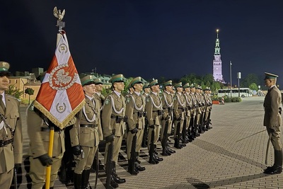 kompania reprezentacyjna sg wraz z pocztem sztandarowym stoi w szeregu, zdjęcie zrobione w nocy,  w tle oświetlona wieża kościoła na jasnej górze